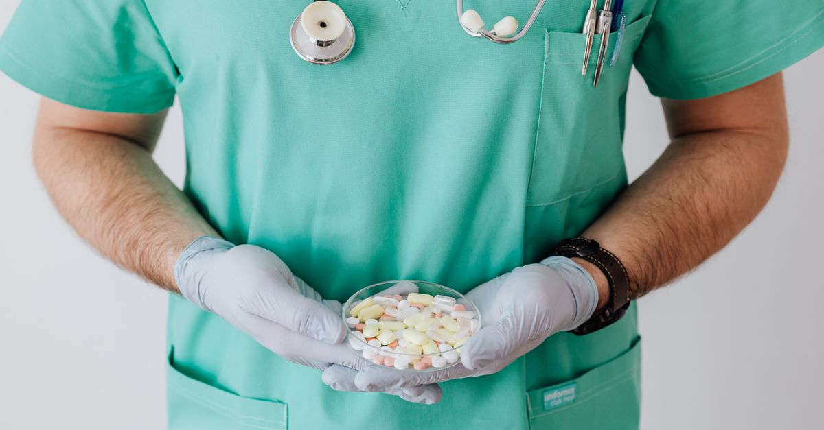 Unrecognizable male doctor wearing medical uniform and latex gloves holding Petri dish filled with assorted drugs while standing near white wall in hospital